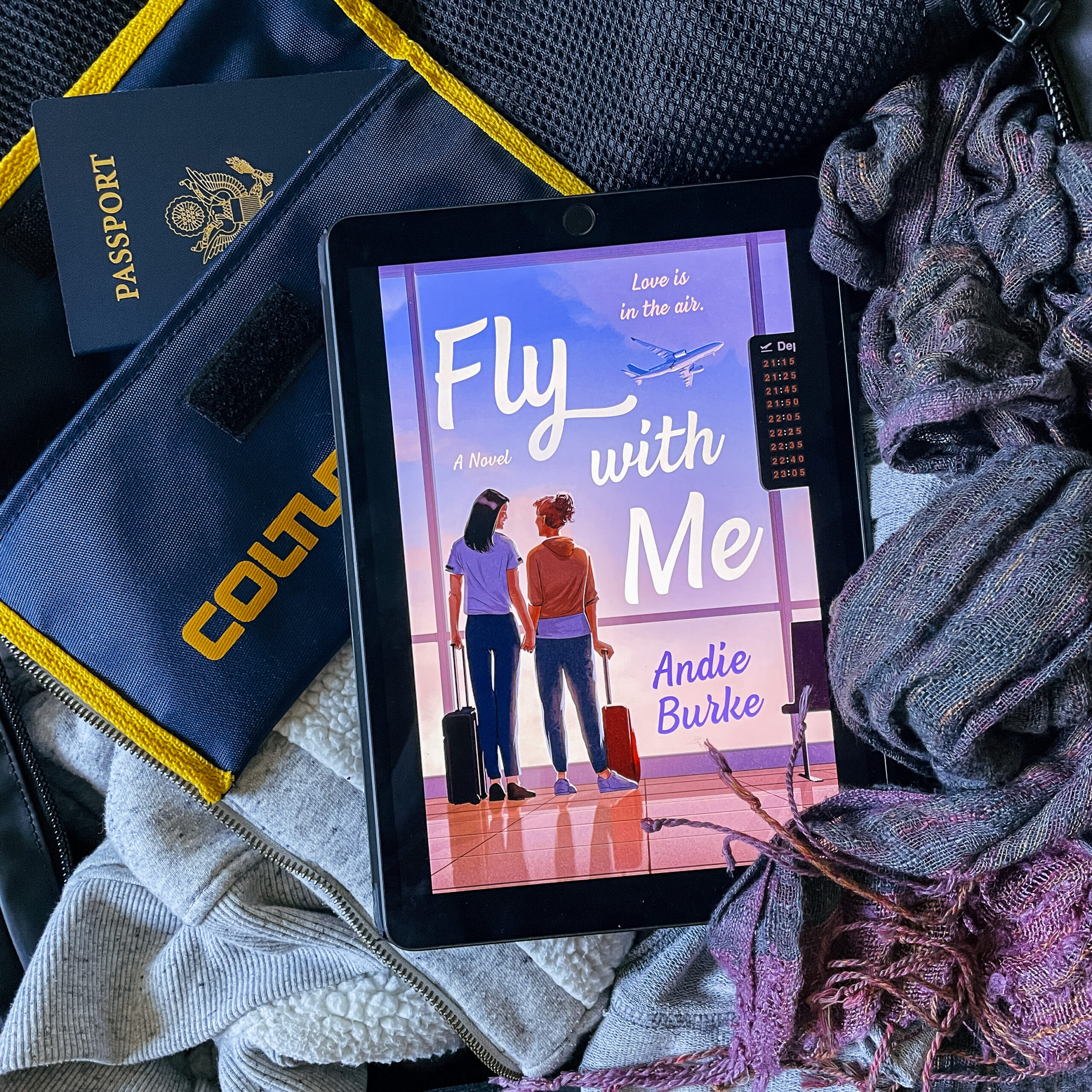 Photo of an ipad with the ebook cover of Fly With Me by Andie Burke displayed on the screen. The ipad is on top of an open suitcase, in which we can see clothes and a passport.